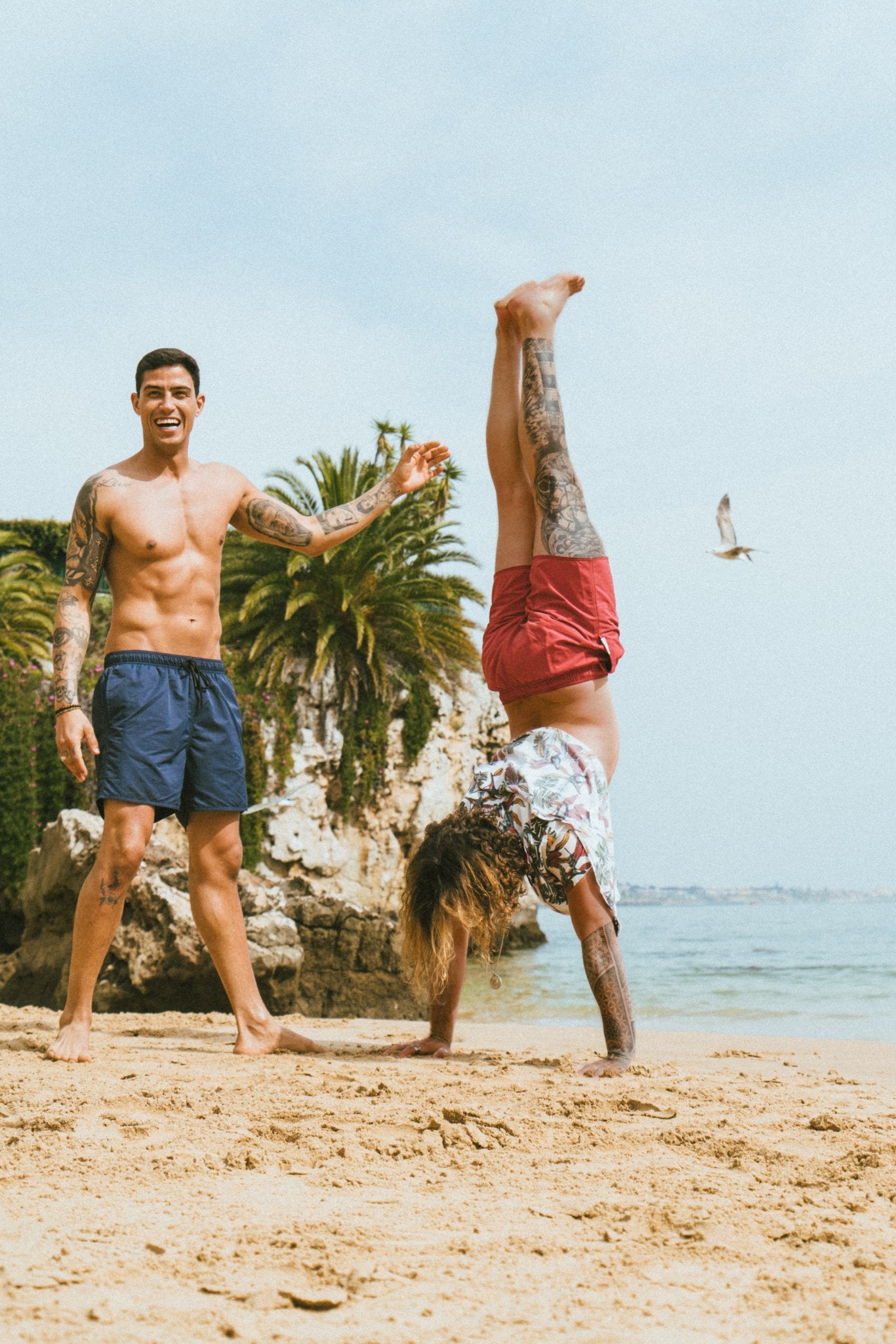 two guys on the beach, one is doing a handstand he is wearing red swim shorts. the other is standing next to it smiling. he is wearing blue swim trunks. the background has the sea, rocks and palmtrees