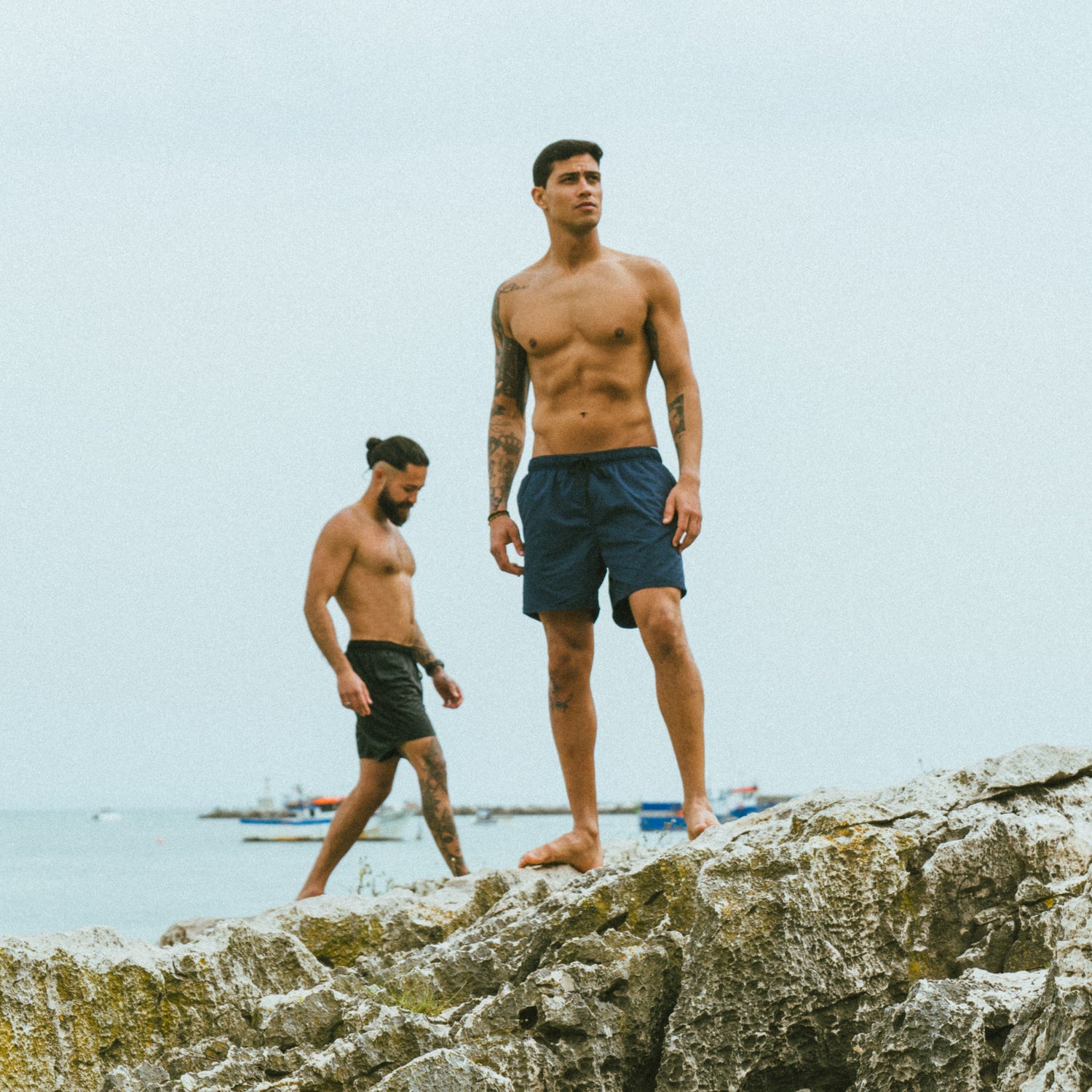 2 models standing on a cliff overlooking the water. The guy in the front is looking serious and wearing navy blue swim trunks. the guy in the back is walking and looking down, he is wearing black swim shorts. the sky is a bit gray