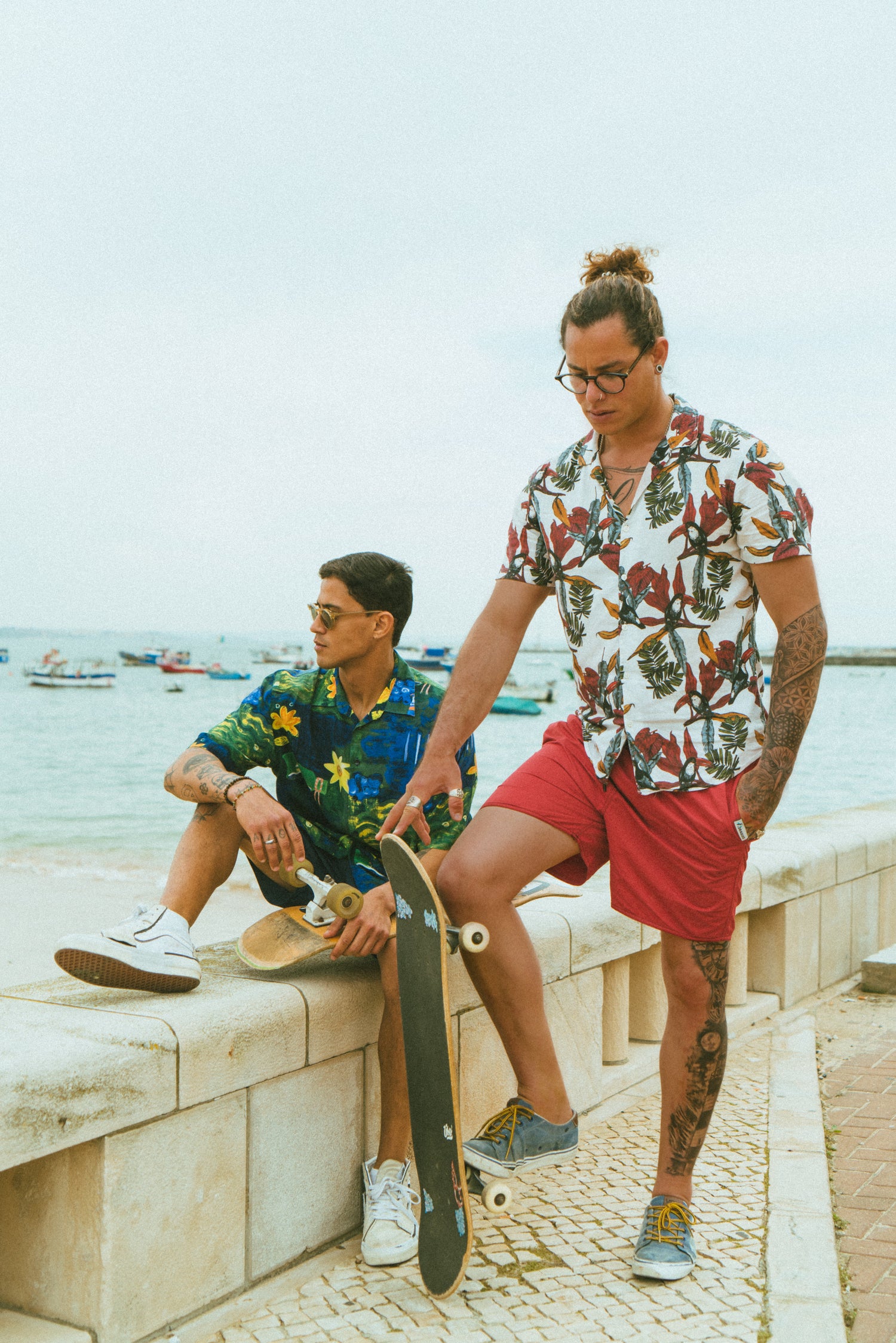 Two models with skateboards, one is sitting on a concrete wall next to the sea wearing the blue swim trunks, the other one is standing next to him wearing the red swim shorts. 