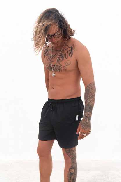 The Cruiser Swim trunks in black from the front, a guy i standing in front of a white wall