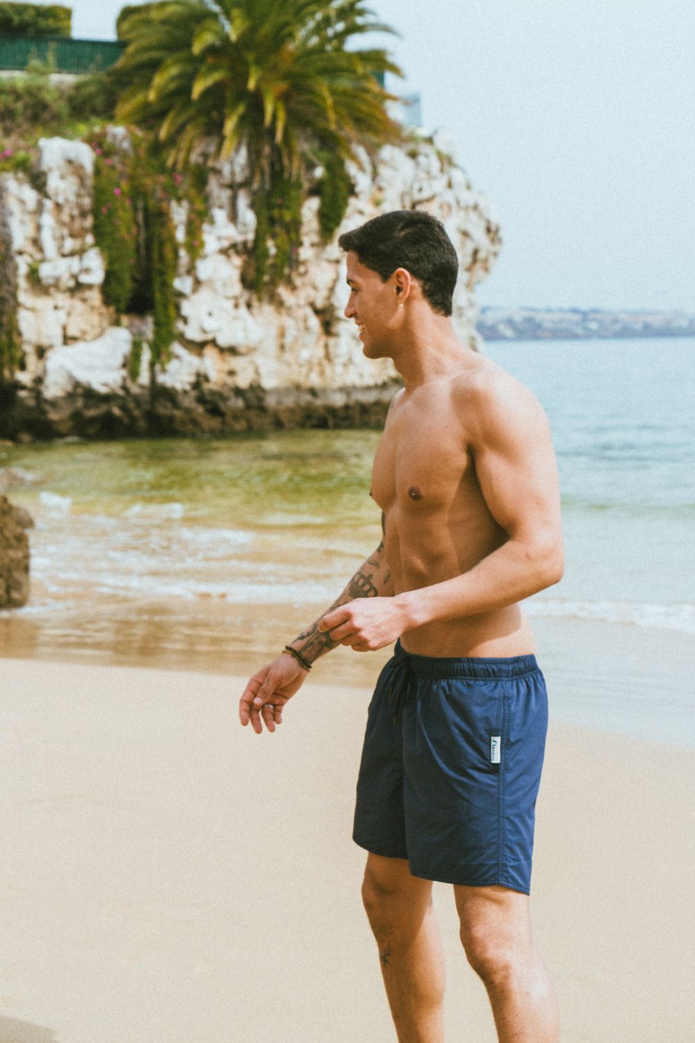 Guy on the beach wearing blue swim trunks. In the background there is the sea, cliffs and palmtrees