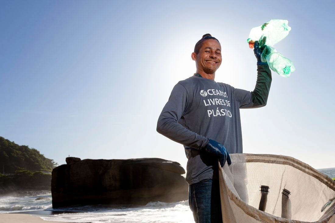 A man collecting ocean plastic for the plastic bank. He is standing on the beach holding a big bag in one hand and in the other hand he holds up 2 plastic bottles. He is smiling